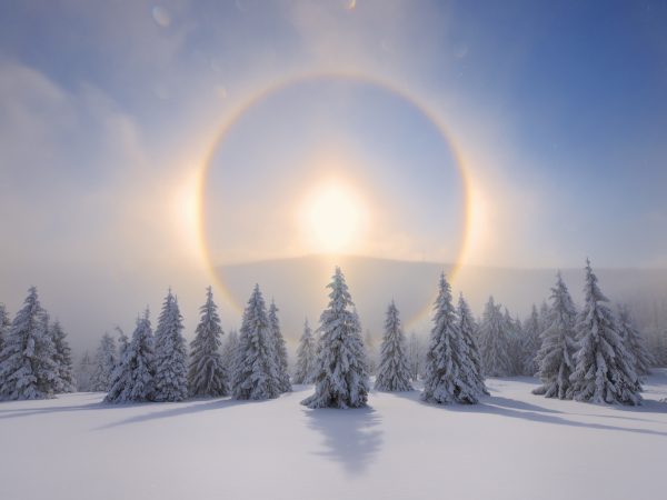 Halo (Icebow or gloriole) around sun with winter landscape. A Halo is an optical phenomenon produced by ice crystals creating colored or white arcs and spots in the sky. The light penonmenon ocurs when ice crystals are in the air reflected the sunlight. This ring can be like  a rainbow. Erzgebirge, Ore Mountains, Fichtelberg, Saxony, Germany.